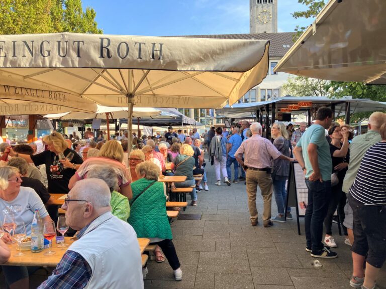Menschen sitzen an Tischen und Bänken gesellig zusammen und geniessen die Atmosphäre des Winzerfestes auf dem Fronhof in Solingen bei abendlicher Stimmung